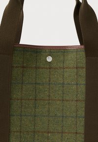 Πράσινο tweed ύφασμα με καφέ και μπλε καρό μοτίβο, καφέ δερμάτινες λεπτομέρειες και φαρδιές σκούρες καφέ υφαντές λαβές σε μια τσάντα.