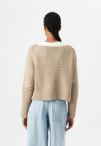 Beige Strickpullover mit offener Maschenstruktur und weitem Ärmel. Der verkürzte Schnitt verfügt über einen weißen Kragen. Kombiniert mit hellblauen weit geschnittenen Jeans.