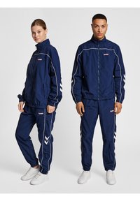 Navy tracksuits made of lightweight fabric, featuring white piping, zip closure, and chevron patterns on sleeves and sides. White sneakers.