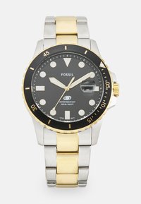Montre pour hommes avec un cadran noir, des accents dorés, un bracelet en métal argenté et doré, une fenêtre de date et étanche jusqu'à 100 mètres.