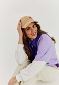 Beżowa czapka z różowym logo, fioletowy i biały kolorowy sweter, kremowe spodnie, modelka z długimi brązowymi włosami, uśmiechająca się, w pozycji siedzącej.