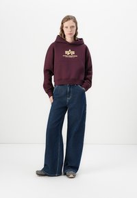 Felpa corta color marrone con un logo dorato sul petto, abbinata a jeans a gamba larga blu scuro e sneakers grigie. Tessuto liscio, design casual.
