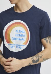 Tamsiai mėlynos spalvos medvilninis marškinėlis su apvalia kreminės spalvos grafika, daugiaspalviais lankais, tekstu "BLEND DENIM ORIGINAL" ir "EST. 1993."