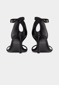 Paire de sandales noires à talons hauts avec brides chevilles et détails découpés, vues de dos sur un fond clair uni.