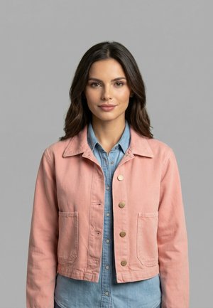 Giovane donna con capelli scuri che indossa una giacca rosa con bottoni sopra una camicia di jeans blu, in piedi davanti a uno sfondo grigio.