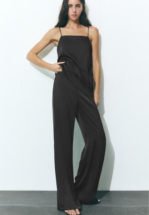 Femme portant un haut en satin noir sans manches et un pantalon à jambes larges, debout contre un mur blanc, une main dans la poche.