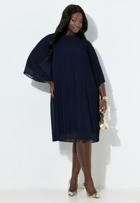 Granatowa plisowana sukienka z dekoltem typu keyhole i szerokimi rękawami. Połączona z przezroczystymi, spiczastymi szpilkami i złotą kopertówką.