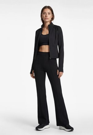 Woman standing wearing a black zip-up jacket, black sports bra, black high-waist flared pants, and black sneakers against a white background.