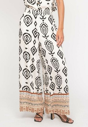 Pantalon large blanc avec motifs géométriques noirs et bordure décorative brune, porté avec des sandales à talons hauts noires à lanières.