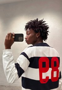 Long-sleeve striped shirt in navy and white with a large red "28" on the back. Material appears smooth with ribbed cuffs. Black smartphone held.