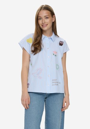 Junge Frau trägt ein blau gestreiftes Kurzarmhemd mit Stickereien von Eule, Blumen, Herzen und dem Text "Vergangenheit, Gegenwart & Zukunft", kombiniert mit blauen Jeans.