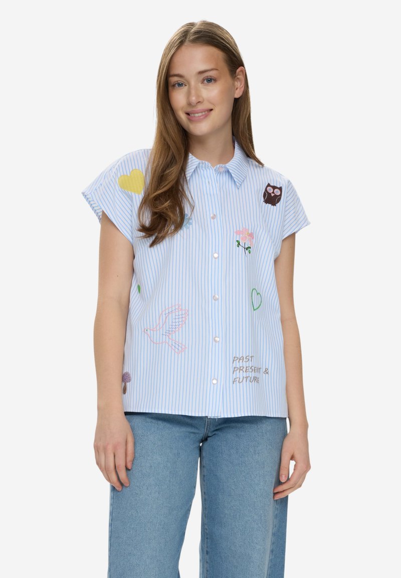 Nuori nainen sinis stripes-pilkullinen lyhythihainen paita, jossa on kirjailtu pöllö, kukkia, sydämiä ja teksti "Past Present & Future", yhdistettynä sinisiin farkkuihin.