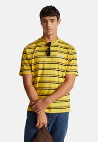 Žluté tričko s černými vodorovnými pruhy, s kulatým výstřihem a krátkými rukávy. Na přední straně je logo "Stranger Things".