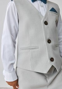 Gilet grigio chiaro con un tessuto texturizzato, caratterizzato da tre bottoni scuri, due tasche frontali e un fazzoletto da taschino blu con motivo. Camicia bianca sotto.