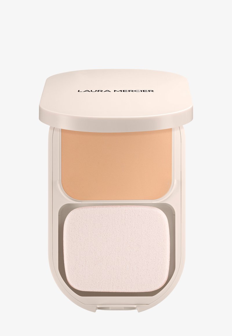 Base en polvo compacto con un tono beige claro en un estuche redondeado de color crema. Incluye una esponja aplicadora suave y blanca en la parte inferior.
