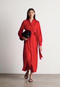 Czerwona satynowa sukienka o kroju kopertowym z długimi rękawami, z kwiatowym akcentem w talii. Modelka trzyma czarną torebkę typu kopertówka o fakturowanej powierzchni.