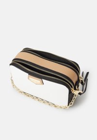Kosmetyczka zapinana na zamek, wykonana z białego, teksturowanego materiału, z beżowymi i czarnymi akcentami, złotymi elementami metalowymi oraz łańcuszkiem na ramię.