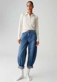 Polo a maniche lunghe color crema a coste e jeans larghi di denim blu con orli arrotolati, indossati con calzini bianchi, su uno sfondo neutro.