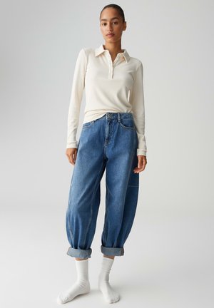 Polo a maniche lunghe color crema a coste e jeans larghi di denim blu con orli arrotolati, indossati con calzini bianchi, su uno sfondo neutro.