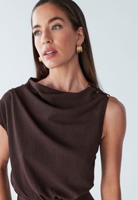 Haut marron sans manches avec un col drapé, tissu texturé et boutons décoratifs dorés sur une épaule.