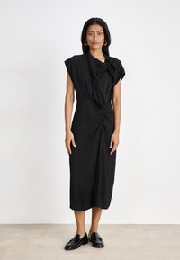 Robe midi noire en tissu doux, avec un col drapé, des manches courtes et un détail froncé à la taille, associée à des chaussures noires brillantes.