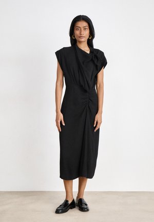 Black midi dress in soft fabric, featuring a draped cowl neck, short sleeves, and gathered detail at the waist, paired with glossy black shoes.