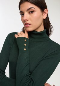 Ciemnozielony sweter z golfem o prążkowanej strukturze i trzech złotych guzikach na mankietach. Dopasowany krój.