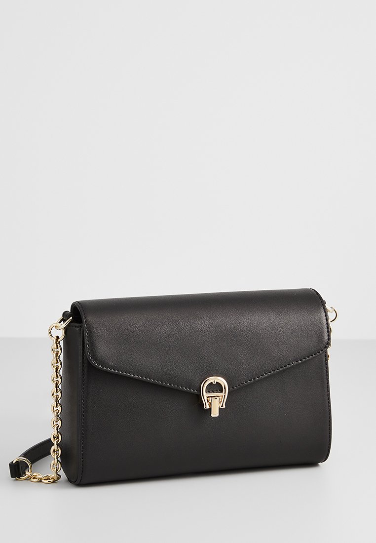 Schwarze Kunstleder-Crossbody-Tasche mit Klappe, goldenen Hardware-Elementen, einem dekorativen Schnallenakzent und einem Kettenriemen. Glatte Textur, rechteckige Form.
