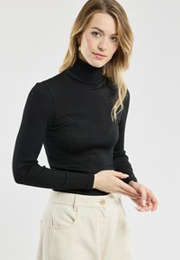 Frau mit langen blonden Haaren, die einen schwarzen Rollkragenpullover und beigefarbene Hochbundhosen trägt, schaut mit zusammengelegten Händen in die Kamera.