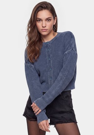 Ung kvinde med langt, bølget hår iført en blå tekstureret cardigan med knapper, sorte slidte shorts og stjerneformede øreringe, der poserer med korslagte arme.