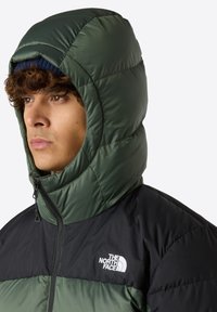 Grön och svart puffjacka med hög krage och huva. Har quiltad yta och framsidans dragkedja. Logotyp visas på bröstet.