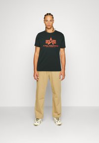 Tricou negru din bumbac cu logo-ul portocaliu "Alpha Industries", asortat cu pantaloni bej și adidași albi. Modelul stă în fața unui fundal neutru.