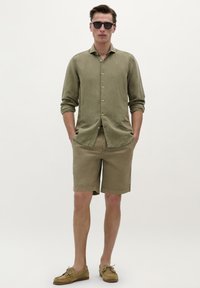 Cămașă verde măslinie cu butoni și mâneci lungi, pantaloni scurți bej și pantofi moccasin de culoare maro deschis; model purtând ochelari de soare negri.