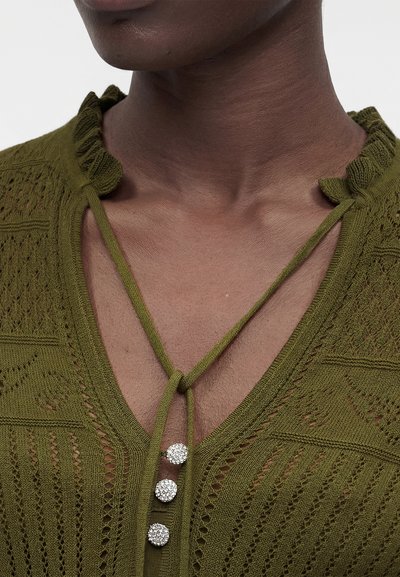 Κοντινό πλάνο σε λαιμό και άνω μέρος στήθους με σκουρόχρωμο δέρμα που φοράει πράσινο στενό πλεκτό τοπ με V-λαιμόκοψη, κορδόνια δέσιμου και τρία στρογγυλά κουμπιά με υφή.