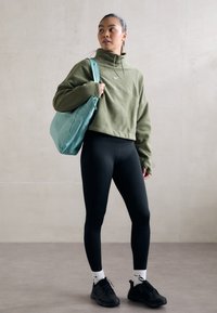 Cappuccio corto verde oliva, leggings neri, scarpe da ginnastica nere, calze bianche con logo, che porta una borsa turchese chiara. Texture lisce.