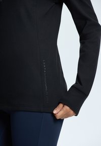 Svart långärmad träningsskjorta med en slät textur, med en logodetalj på sidan. Kombinerad med marinblå tights som har en åtsittande design.