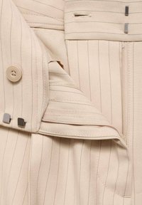 Veste beige sur mesure présentant de fines rayures verticales, fermeture à boutons, fermeture éclair et accents métalliques. Tissu doux avec un design structuré.