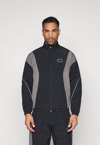 Mann in einer schwarz-grauen Trainingsjacke mit Reißverschluss und passenden Hosen, stehend vor einem schlichten hellen Hintergrund.