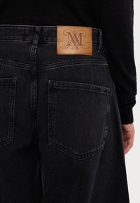 Mustat farkut nahkapaikalla, jossa on kohokuvioitu logo, viisi taskua ja suoransuuntaiset lahkeet. Raskaspintainen kangas, ei näkyviä kuvioita.