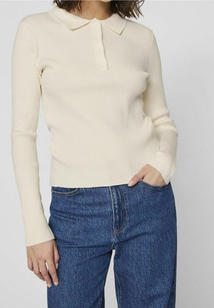 Femme portant un pull polo à manches longues côtelé crème rentré dans un jean bleu taille haute, main reposant dans la poche.