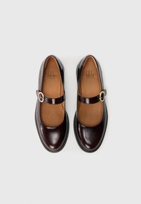 Zapatos de cuero marrón con punta redondeada, una correa decorativa con un detalle de hebilla y una suela de goma texturizada. El interior presenta un forro de cuero beige.