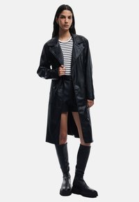 Czarny płaszcz trench z sztucznej skóry z szerokimi klapami, zapięciem na guziki i bocznymi rozcięciami, noszony na czarno-biały paskowany top oraz krótkie spodenki, zestawiony z kozakami za kolano.