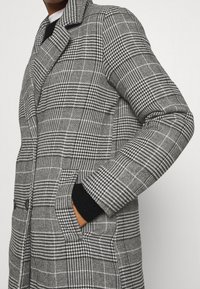 Persona vestida con un abrigo de patrón de espiga blanco y negro, con bolsillos frontales y botones, con la mano metida en uno de los bolsillos.