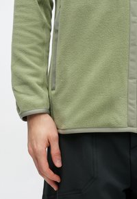 Prim-plan al unei mâini care se odihnește lângă o jachetă de fleece verde deschis și pantaloni negri cu cusături vizibile și detalii ale buzunarelor.