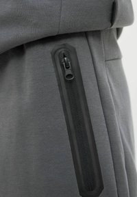 Un jogging en tissu gris présente une vue en gros plan d'une poche zippée noire avec un bord texturé noir, mettant en valeur un design élégant et des détails fonctionnels.