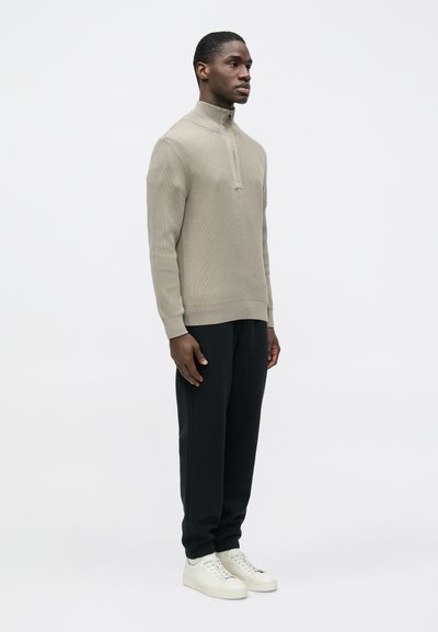 Pull gris clair côtelé avec un col montant, associé à un pantalon noir et des baskets blanches. Le tissu semble texturé et confortable.