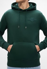 Hoodie verde scuro realizzato in morbido tessuto, con tasca frontale, coulisse regolabili e logo in rilievo sul petto.
