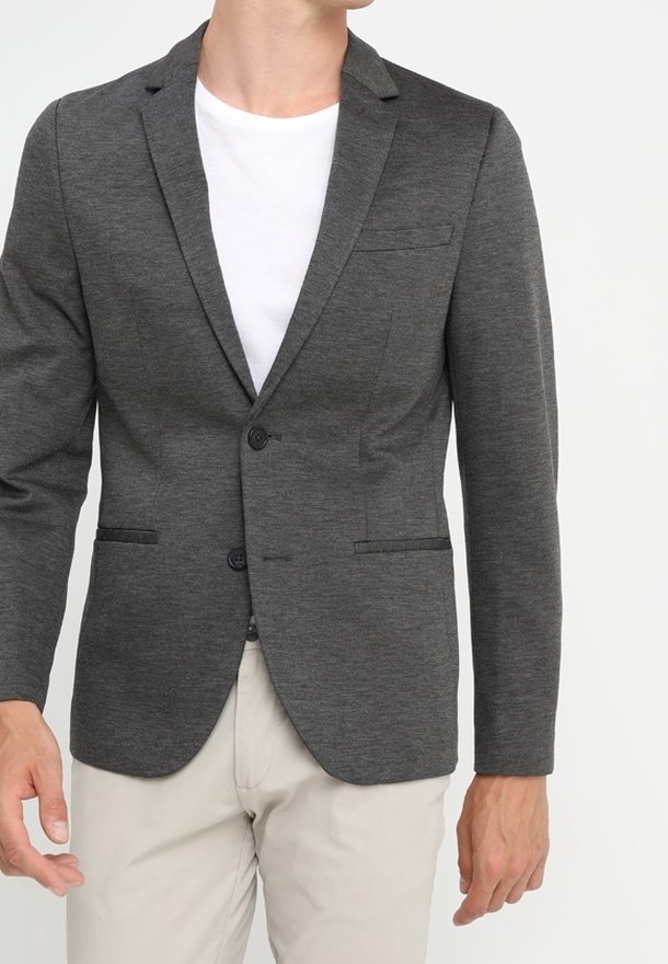 Uomo che indossa un blazer aderente grigio scuro sopra una camicia bianca e pantaloni beige chiaro, in piedi contro uno sfondo semplice.