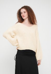 Εκρού, υπερμεγέθη πλεκτό πουλόβερ με μεγάλο V-λαιμόκοψη και ριμπ μανσέτες, συνδυασμένο με μαύρη, ρυτιδωτή φούστα. Μαλακό, υφή ύφασμα.