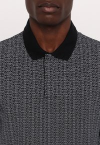 Swarfarget poloskjorte med svart og hvitt mønster, solid svart krage, geometrisk design og knappestolpe ved halsen.